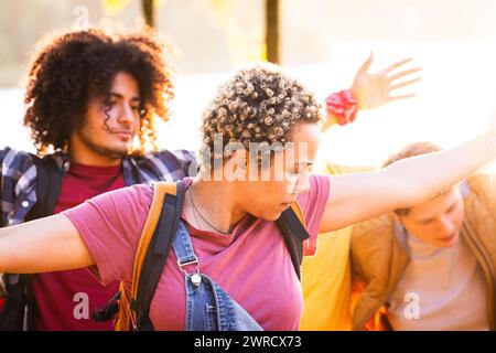 Dieses Bild zeigt eine Gruppe junger Freunde in einem Moment der Entspannung und Interaktion während eines Outdoor-Abenteuers. Das von der Sonne hinterleuchtete Foto fängt die intime Dynamik der Gruppe ein. Eine junge Frau mit kurzen lockigen Haaren sieht man, wie sie ihren Arm dehnt, vielleicht in einem Moment des Gesprächs oder einer Dehnung, während ein junger Mann mit lockigen Haaren anschaut. Der weiche Fokus auf dem Hintergrund lässt vermuten, dass ihre Umgebung in das goldene Licht des späten Nachmittags getaucht ist. Junge Freunde, die ein sonnendurchflutetes Outdoor-Abenteuer genießen. Hochwertige Fotos Stockfoto