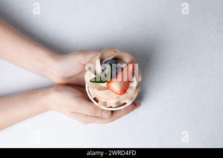 Blick von oben auf die Kinderhände, die Schokoladen-Cupcake mit Frischkäse-Zuckerguss halten, Obst auf Betonhintergrund Stockfoto