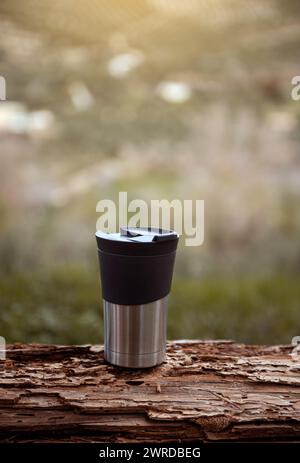 Vertikale Aufnahme eines Reisebechers auf einem Baumstamm verschwommenen Naturhintergrund mit Werbespot. Eine Edelstahltasse mit heißem Getränk, Kaffee oder Tee für Stockfoto