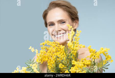 Frau mittleren Alters, die Eleganz mit Mimosa-Bouquet auf blauem Hintergrund präsentiert Stockfoto