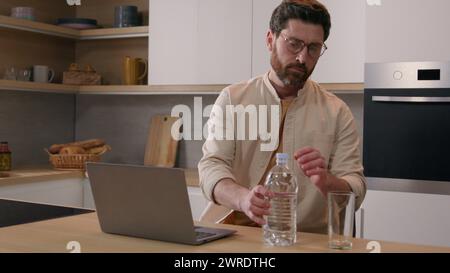Kaukasischer Mann freischaffender Geschäftsmann Benutzer mit Laptop arbeiten von zu Hause entfernt Internet Computer Arbeit in der Küche gießen Wasser von der Flasche ins Glas Stockfoto