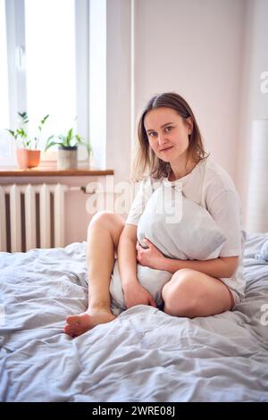 Eine schöne junge Frau in weißem T-Shirt auf dem Bett, das sich an das Kissen schmiegt Stockfoto