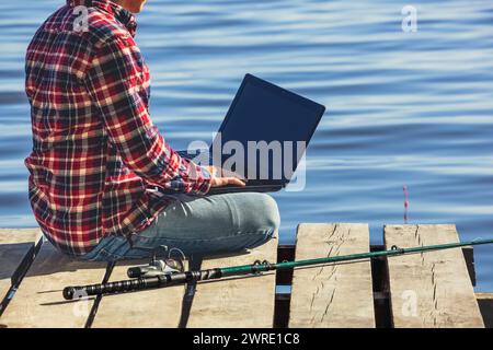 Ein Fischer arbeitet an einem Laptop, sitzt auf einem hölzernen Pier in der Nähe des Sees, daneben befindet sich eine Angelstange. Stockfoto