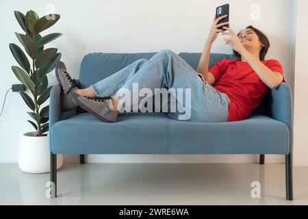 Eine schöne Frau in einem roten Hemd, die auf dem Sofa liegt und mit ihrem Telefon spielt Surfen Sie in entspannter, glücklicher Stimmung im Internet. Stockfoto