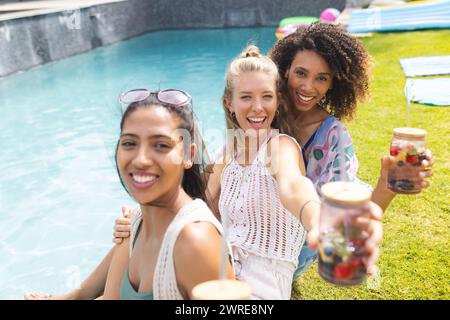 Verschiedene Freundinnen genießen ein sonniges Treffen am Pool und halten erfrischende Getränke Stockfoto