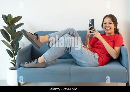 Eine schöne Frau in einem roten Hemd, die auf dem Sofa liegt und mit ihrem Telefon spielt Surfen Sie in entspannter, glücklicher Stimmung im Internet. Stockfoto