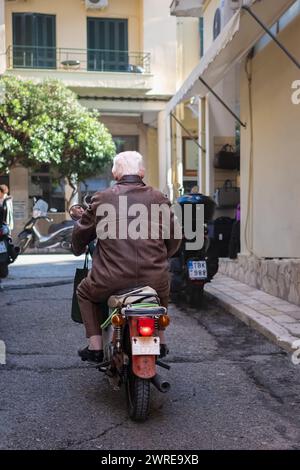 Senior Gentleman, der ein Oldtimer-Motorrad auf der Straße einer Altstadt in Griechenland fährt. Älterer Mann, Rentner, der auf Korfu Fahrrad fährt. Reisefoto, aktiv Stockfoto
