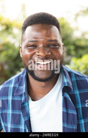 Ein afroamerikanischer Mann lächelt in einem Garten und zeigt einen kurzen Bart und ein blaues kariertes Hemd Stockfoto