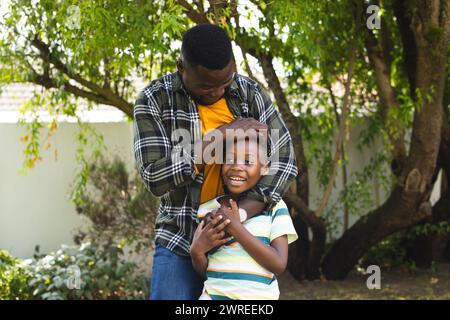 Der afroamerikanische Vater umarmt einen fröhlichen Sohn in einem sonnigen Garten Stockfoto