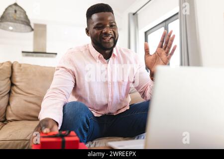 Ein afroamerikanischer Mann in einem rosa Hemd winkt bei einem Videoanruf an einem Laptop-Bildschirm und hält ein rotes Geschenk Stockfoto