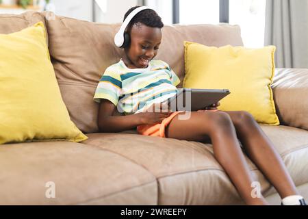 Der afroamerikanische Junge genießt ein Tablet, während er Kopfhörer auf dem Sofa trägt Stockfoto