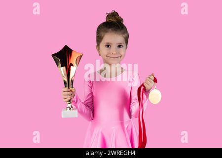 Fröhliches Sportmädchen, das den Sieg auf rosa Hintergrund feiert, trägt Goldmedaille und zeigt die Trophäe, die sie gewonnen hat. Hochwertige Fotos Stockfoto