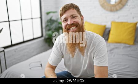 Wachen Sie auf zu einem charmanten Morgen mit einem jungen, glücklichen rothaarigen Mann, der bequem im Bett sitzt, selbstbewusst in seinem gemütlichen Schlafzimmer lächelt und sich entspannt Stockfoto