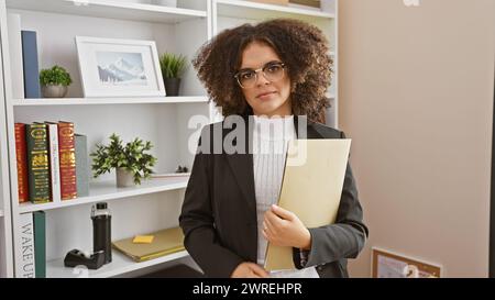 Eine junge hispanische Frau mit lockigem Haar, die professionell gekleidet ist, hält eine Mappe in einem Büro und strahlt Selbstvertrauen aus. Stockfoto