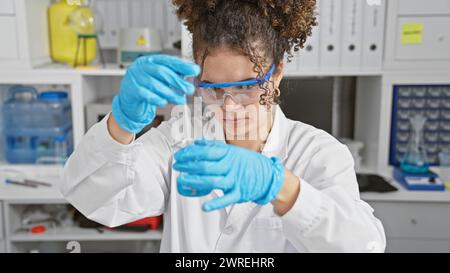 Eine junge erwachsene hispanische Frau mit lockigem Haar führt ein Experiment in einem Labor durch und trägt Schutzbrille und Handschuhe. Stockfoto