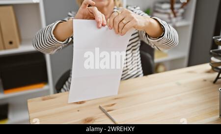 Eine Frau mittleren Alters zerreißt in einem Büro ein weißes Papier und zeigt eine Mischung aus Frustration und Entschlossenheit. Stockfoto