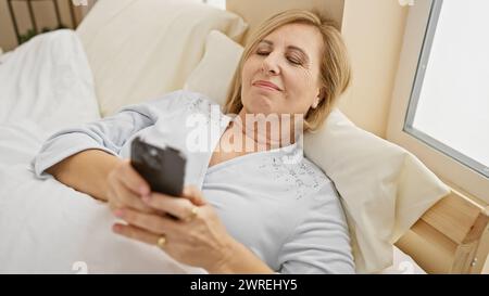 Eine ruhige Frau mittleren Alters entspannt sich im Bett mit einem Smartphone in einem gemütlichen Schlafzimmer und verkörpert Freizeit und moderne Konnektivität. Stockfoto