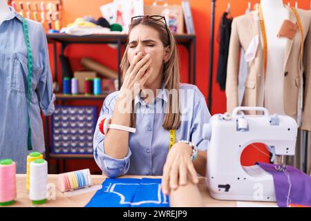 Hispanische junge Schneiderin-Designerin im Atelierzimmer gelangweilt, müde zu gähnen, den Mund mit der Hand zu bedecken. Ruhelos und Schläfrigkeit. Stockfoto