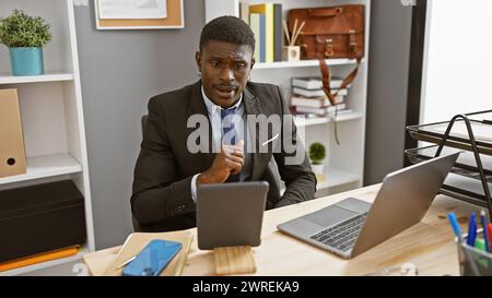 Ein professioneller afroamerikanischer Mann im Anzug saß nachdenklich an seinem Schreibtisch mit Laptop und Tablet. Stockfoto