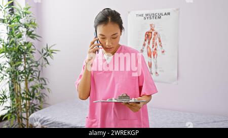 Eine junge asiatische Frau in einem rosafarbenen Gewand, die an einem Telefon mit einem Klemmbrett in einem Klinikinnenraum spricht. Stockfoto