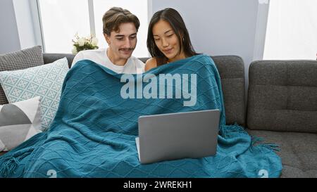 Interrassisches Paar, das sich einen gemütlichen Moment unter einer Decke auf der Couch mit einem Laptop in einem modernen Innenraum teilt. Stockfoto