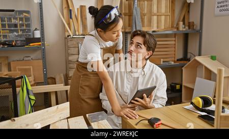 Mann und Mädchen in Schürzen arbeiten in einer Holzwerkstatt mit digitalen Tablets und Werkzeugen zusammen. Stockfoto