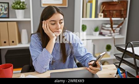 Hispanische Frau mit Smartphone im modernen Büro, die nachdenklich aussieht. Stockfoto
