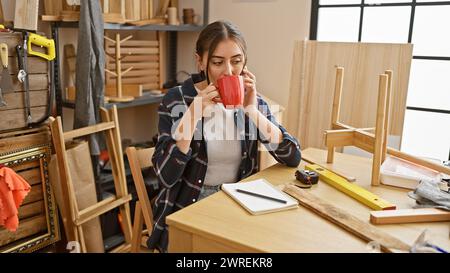 Eine junge hispanische Frau genießt eine Kaffeepause in einer Holzwerkstatt, umgeben von Werkzeugen und Holzstücken. Stockfoto