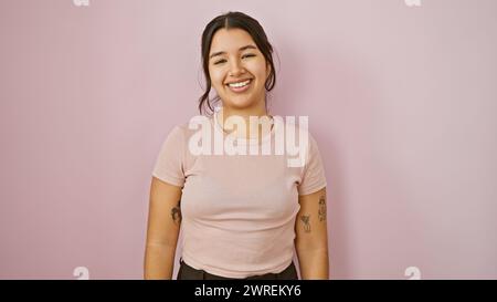 Eine lächelnde junge hispanische Frau posiert lässig vor einem schlichten rosafarbenen Hintergrund und strahlt Selbstvertrauen und Schönheit aus. Stockfoto