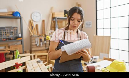 Eine junge hispanische Frau überprüfte Pläne in einer gut ausgestatteten Tischlerei, was auf eine praktische kreative Beschäftigung hinweist. Stockfoto