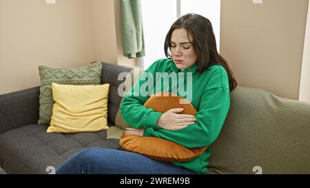 Eine junge kaukasische Frau mit schmerzhaftem Ausdruck sitzt drinnen auf einem grauen Sofa, hält sich an den Bauch und umschließt ein Kissen. Stockfoto