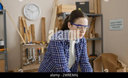 Fokussierte junge Frau mit Schutzbrille in einer Tischlerei, umgeben von Werkzeugen und Holz. Stockfoto