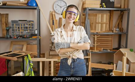 Lächelnde junge kaukasische Frau mit gekreuzten Armen in Schutzkleidung in einer Tischlerei. Stockfoto