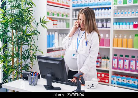 Junge Schwangerenapothekerin, die in der Apotheke am Smartphone mit dem Computer spricht Stockfoto