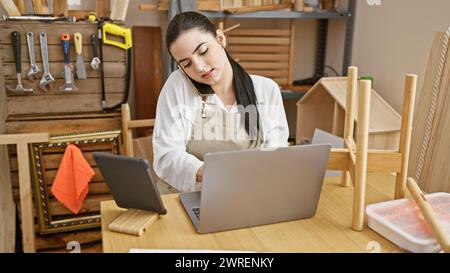 Eine junge Tischlerin, die in einem Telefongespräch arbeitet, während sie in ihrem Holzwerkerstudio arbeitet. Stockfoto