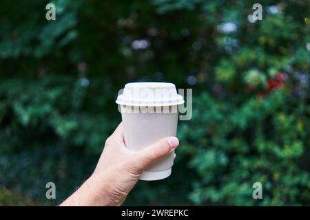 Mann hält draußen eine Einweg-Kaffeetasse mit grünem Laub im Hintergrund. Stockfoto