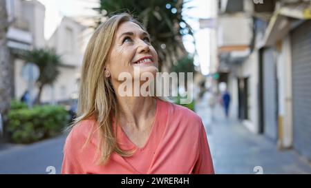 Eine lächelnde blonde Frau mittleren Alters blickt auf, während sie draußen auf einer Stadtstraße spaziert und ein Gefühl von Freude und Zufriedenheit ausstrahlt. Stockfoto