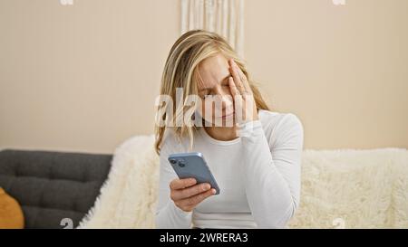 Eine beunruhigte junge Frau in einem Wohnzimmer, die ihr Handy mit Kopfschmerzen ansieht Stockfoto