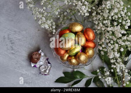 Schokoladen-Ostereier in Goldfolie gewickelt. Weiße Frühlingsblumen und Süßigkeiten von Ester von oben. Strahlendes Stillleben im Frühling. Stockfoto