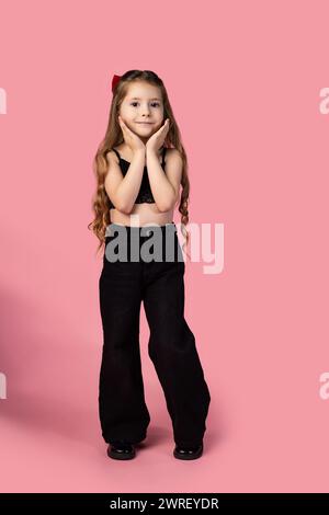 Ein Foto in voller Länge von einem kleinen Mädchen, das mit den Händen unter dem Kinn steht und schwarz gekleidet ist und das Kamerafoto auf einem rosa Hintergrund ansieht. Hallo Stockfoto