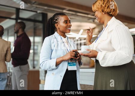 Zwei professionelle afrikanische Geschäftsfrauen führen ein Gespräch auf einer Geschäftskonferenz. Sie halten Kaffeetassen und tragen Konferenzabzeichen, Smili Stockfoto