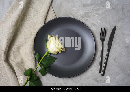 Tischdekoration mit weißer Rose, schwarzem Teller und Besteck auf strukturiertem grauen Hintergrund. Draufsicht, flach. Stockfoto