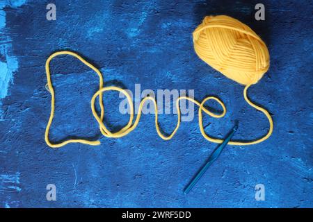 Word Love aus gelbem Wollprofil auf blau strukturiertem Hintergrund mit Kopierraum. Foto von oben mit Wollgarn und Häkelnadel. Hobby und Freizeit Stockfoto