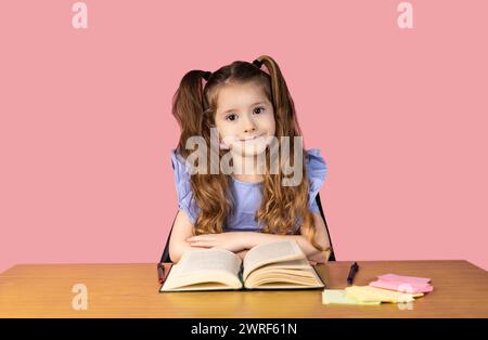 Ein cleveres und aufgeräumtes Mädchen mit zwei Pferdeschwänzen hält ihre Hände auf der Bank und sieht lächelnd auf die Kamera auf einem rosa Hintergrund aus. Konzept von Stockfoto