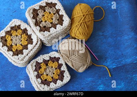 Gehäkelte Oma-Quadrate aus naturbrauner, senfeiner und beiger Wolle. Weiches und flauschiges Häkelschmuck auf blau strukturiertem Hintergrund. Stockfoto