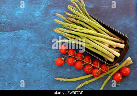 Frischer grüner Spargel und Kirschtomaten auf blau strukturiertem Hintergrund mit Kopierraum. Gesunde Zubereitung des Abendessens. Ausgewogenes Ernährungskonzept. Stockfoto