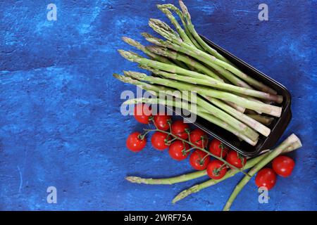 Frischer grüner Spargel und Kirschtomaten auf blau strukturiertem Hintergrund mit Kopierraum. Gesunde Zubereitung des Abendessens. Ausgewogenes Ernährungskonzept. Stockfoto