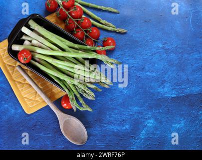 Frischer grüner Spargel und Kirschtomaten auf blau strukturiertem Hintergrund mit Kopierraum. Gesunde Zubereitung des Abendessens. Ausgewogenes Ernährungskonzept. Stockfoto