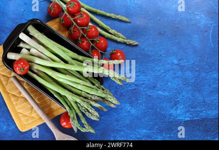 Frischer grüner Spargel und Kirschtomaten auf blau strukturiertem Hintergrund mit Kopierraum. Gesunde Zubereitung des Abendessens. Ausgewogenes Ernährungskonzept. Stockfoto
