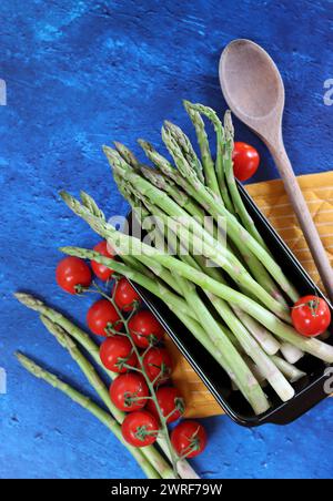 Frischer grüner Spargel und Kirschtomaten auf blau strukturiertem Hintergrund mit Kopierraum. Gesunde Zubereitung des Abendessens. Ausgewogenes Ernährungskonzept. Stockfoto